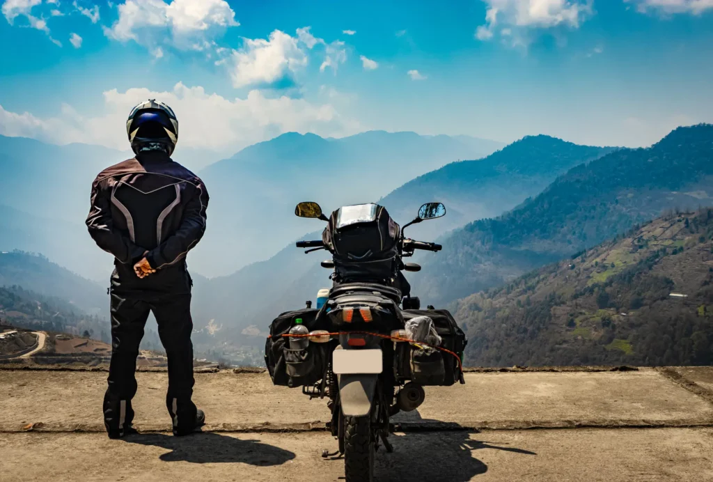 Motorcyclist in gear pauses at scenic overlook with motorcycle, enjoying mountain vista.